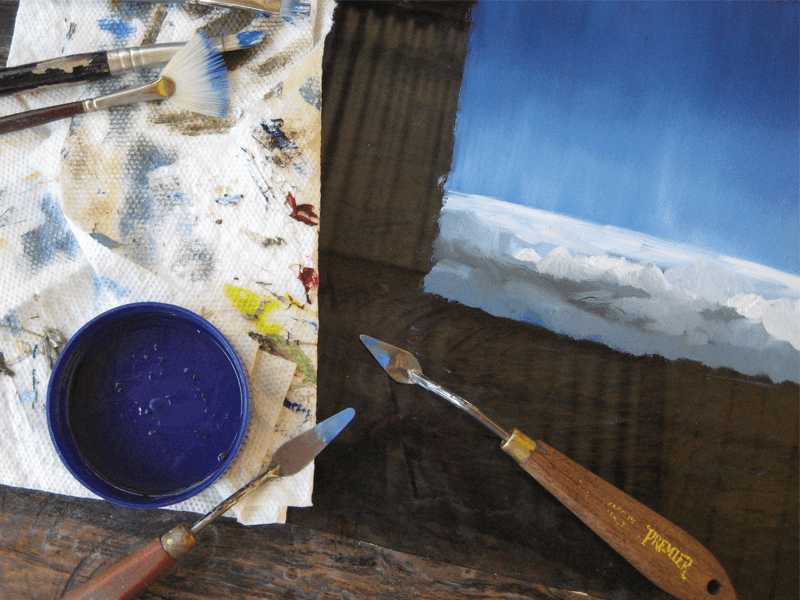 Work in progress: a pot of blue paint and a palette knife beside a glass painting panel.