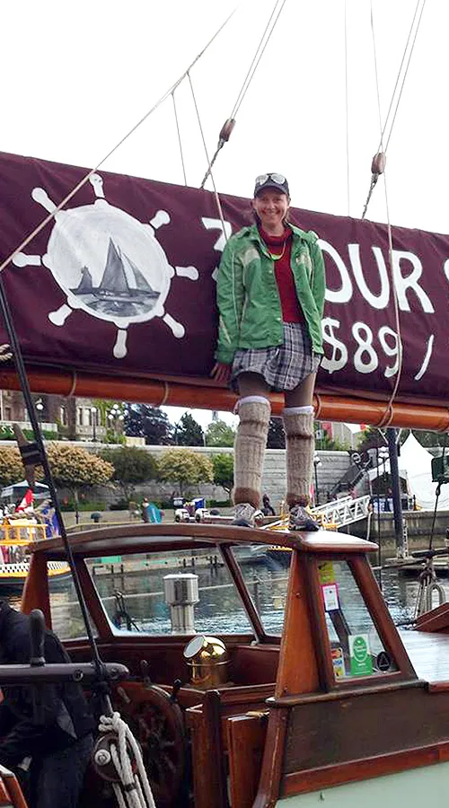 Nic Wood standing on the bowsprit of a tall ship at dock, wearing a green jacket and knitted leg warmers.